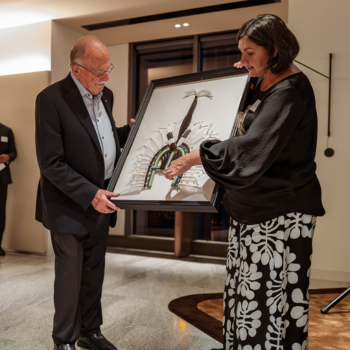 Two people presenting a framed Indigenous artwork at a formal event.